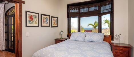 A bedroom with a bed, a nightstand, a desk, a ceiling fan, a window with a view, and framed artwork on the wall.