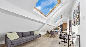 A modern loft bedroom with skylights, a gray sofa, a desk with chairs, and artwork on the walls. - Flat -12 (London)