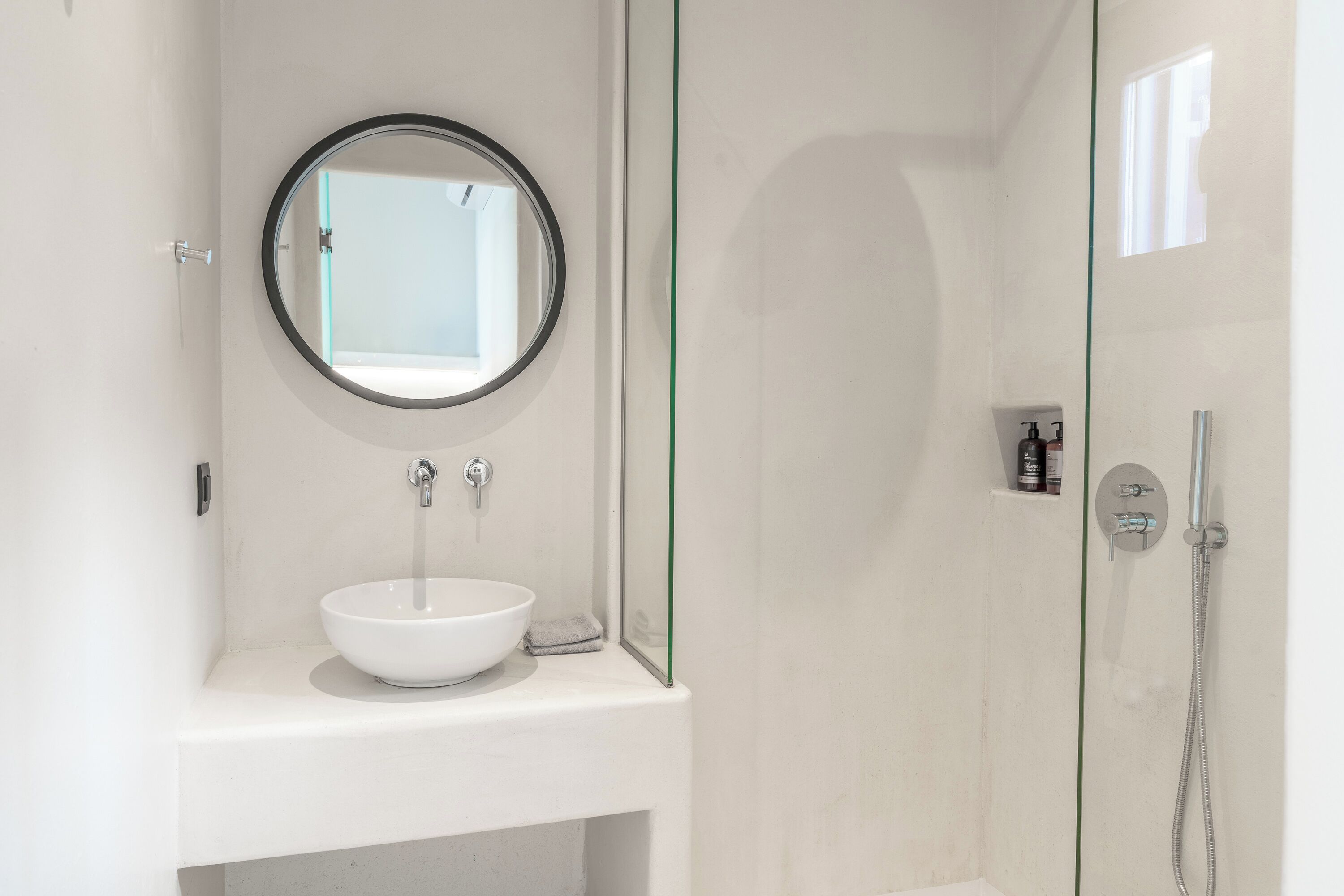 Un baño moderno con un espejo redondo, un lavabo blanco y una ducha con mampara de cristal.