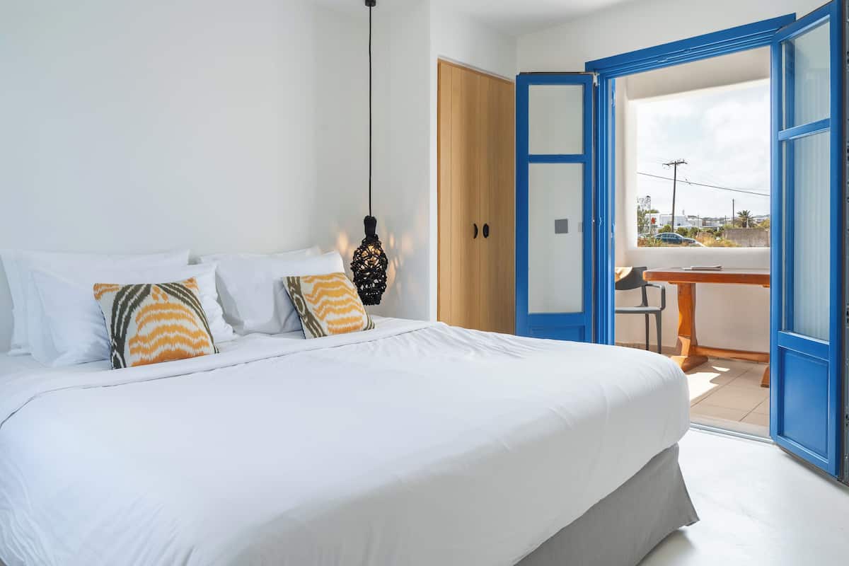 Une chambre à coucher avec un lit, des oreillers, un luminaire suspendu, une porte en bois et une vue sur l’extérieur, visible à travers une porte ouverte.