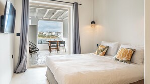 Une chambre à coucher comprenant un lit, un téléviseur, un coin repas avec des chaises et une table, et une vue sur l’extérieur, visible à travers l’encadrement d’une porte.