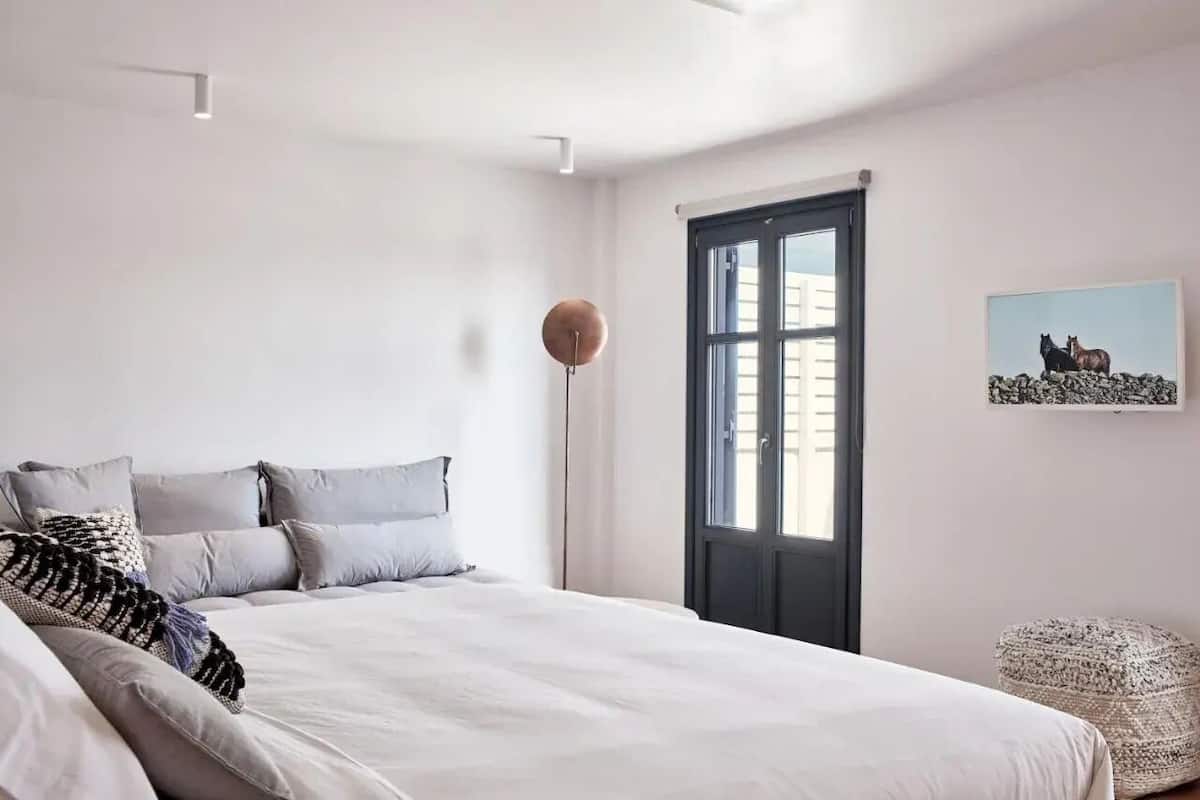 A bedroom with a bed, pillows, a floor lamp, a window, and a wall-mounted picture.
