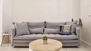 Un canapé gris agrémenté de plusieurs coussins, une petite table ronde en bois sur laquelle est posée une plante, et un mur blanc orné d’un cadre.