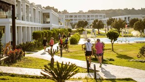 A pathway with people walking, surrounded by greenery and a building with columns. - Levante Athletic Resort (Rhodes)