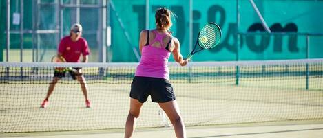 Een tennisbaan met spelers in actie, een net en een groene muur op de achtergrond.