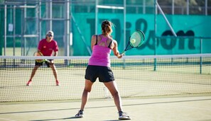A tennis court with players in action, a net, and a green wall in the background. - Levante Athletic Resort (Rhodes)
