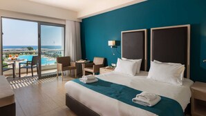 A hotel room with a large bed, two nightstands, a desk, and a chair. There is a view of a pool and buildings through a large window. - Levante Athletic Resort (Rhodes)