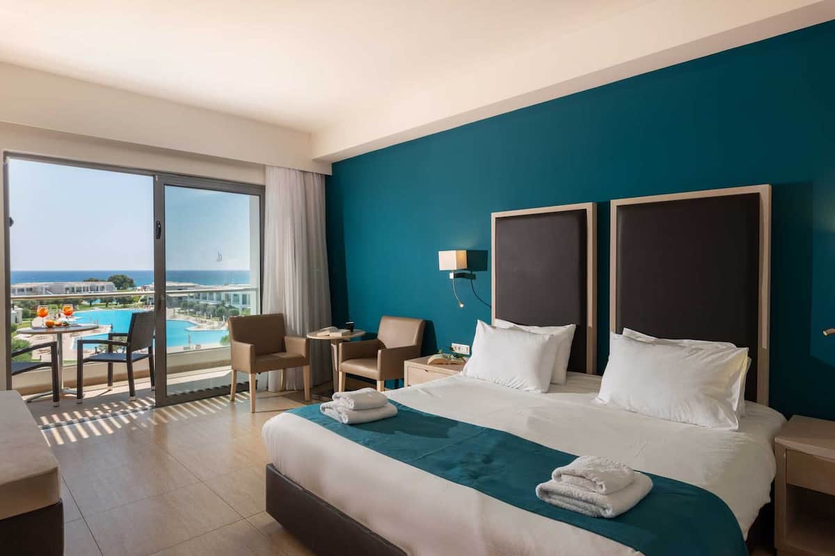 A hotel room with a large bed, two nightstands, a desk, and a chair. There is a view of a pool and buildings through a large window.