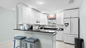 Une cuisine moderne avec des meubles blancs, un îlot central doté de tabourets, un réfrigérateur et un four à micro-ondes.