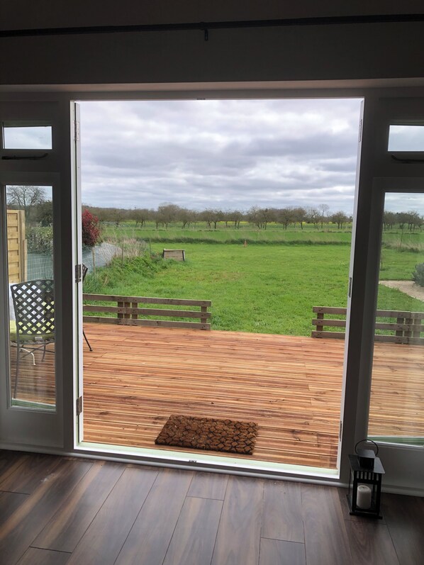 A room with a large glass door leading to an outdoor deck with a wooden floor and a view of a green field.