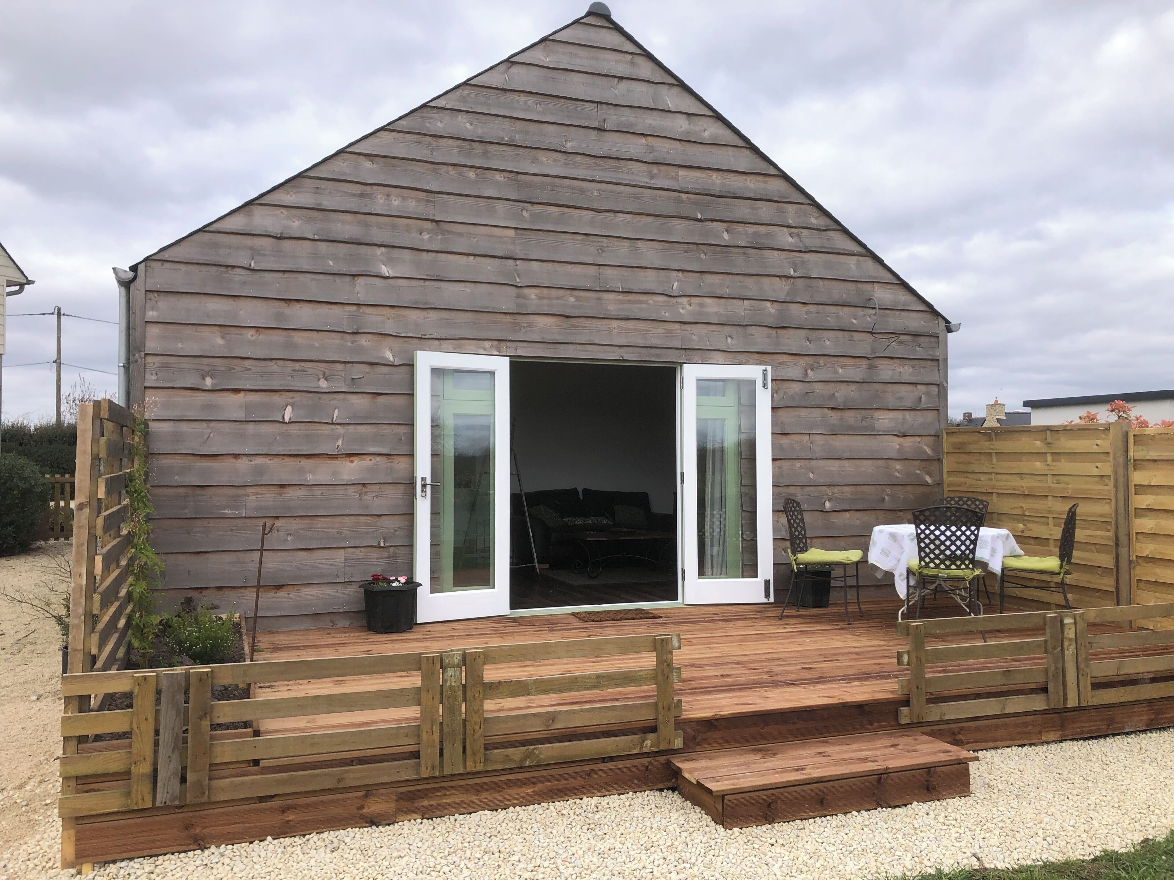 Une cabane en bois avec un toit en pente, une porte-fenêtre coulissante, un petit espace extérieur aménagé avec une table et des chaises, et une allée en gravier.