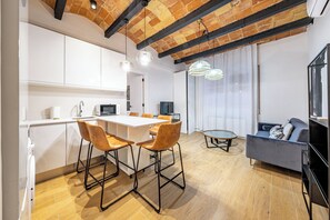 Eine moderne Küche mit weißen Schränken, ein Essbereich mit Holzsesseln, einem runden Couchtisch und einem grauen Sofa.