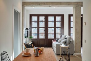Una sala de estar moderna con un sofá, una mesa de comedor con una cafetera y pasteles, y vistas a través de amplios ventanales.