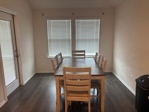 Een eetkamer met een rechthoekige tafel, zes houten stoelen, twee ramen met jaloezieën en een deur.