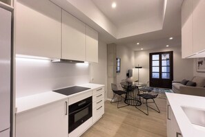 Una cocina moderna con armarios blancos, una placa de inducción negra y un horno empotrado. Hay una zona de comedor con una mesa redonda y sillas, y un sofá en la sala de estar.