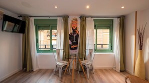 Ein Zimmer mit Glastisch und vier Stühlen, zwei Fenstern mit grünen Rahmen, einem Flachbildfernseher an der Wand und einer Vase mit Zweigen auf der rechten Seite.