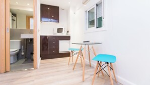 A compact kitchen with a microwave, cabinets, and a window. There is a small round table with two blue chairs. - Ample Bolsa estándar tipo 1 (Barcelona)