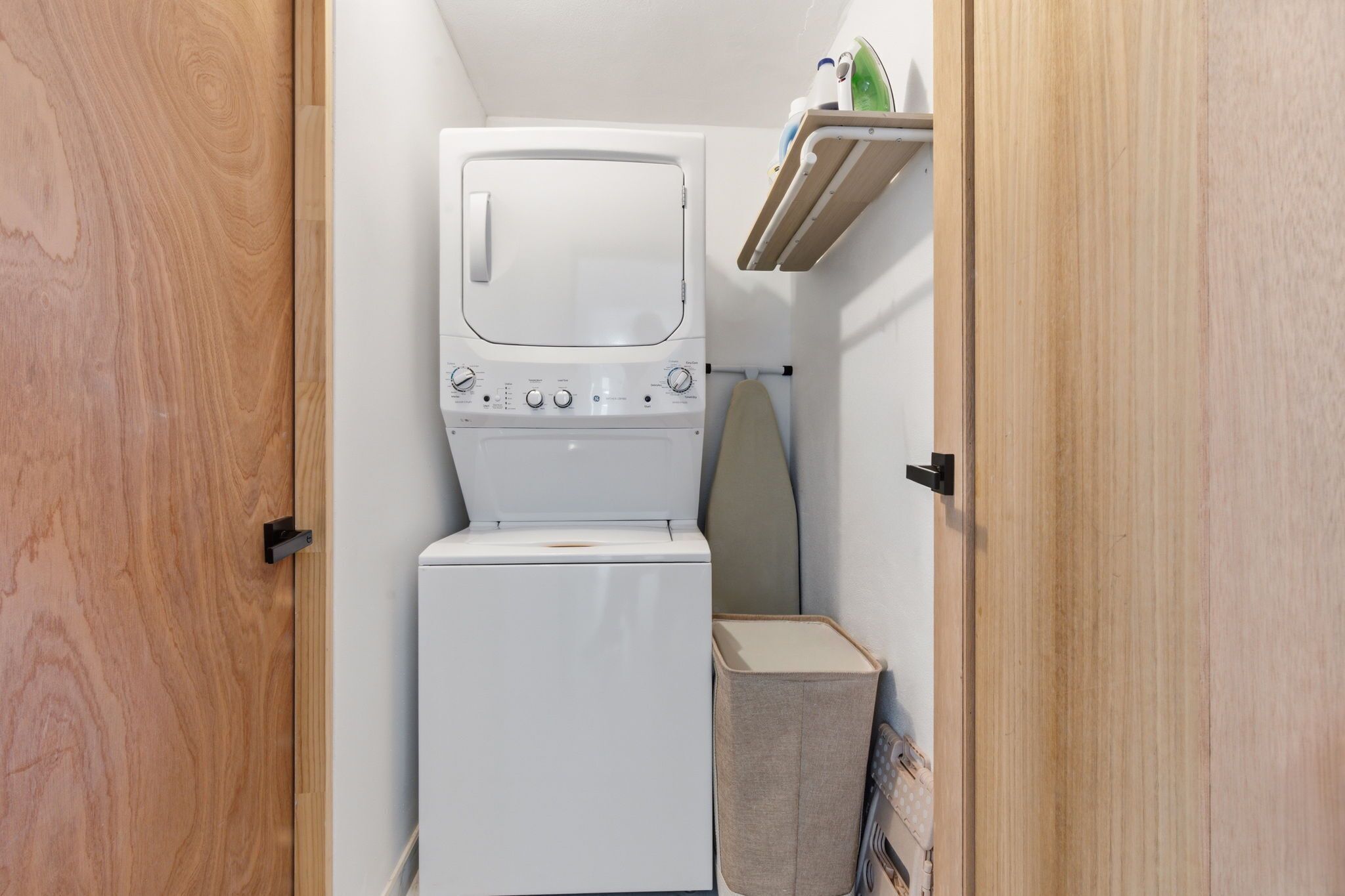 A compact laundry room with a washer and dryer, a small ironing board, and a shelf with toiletries.