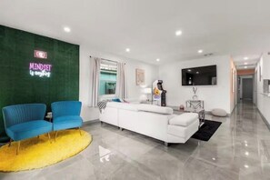 Ein modernes Wohnzimmer mit einem weißen Sofa, blauen Sesseln, einem gelben Teppich, einem an der Wand montierten Fernseher und einem Neonlicht.