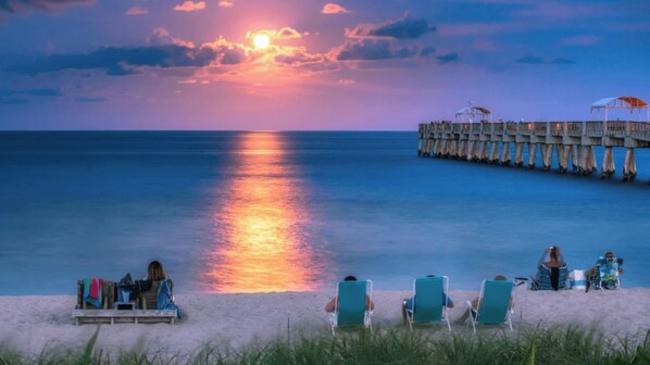 A beach with a pier extending into the water, people relaxing on the sand, and a sunset in the background.