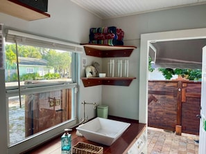A modern kitchen with a large window, a white sink, wooden shelves, and a view of a house and greenery outside.