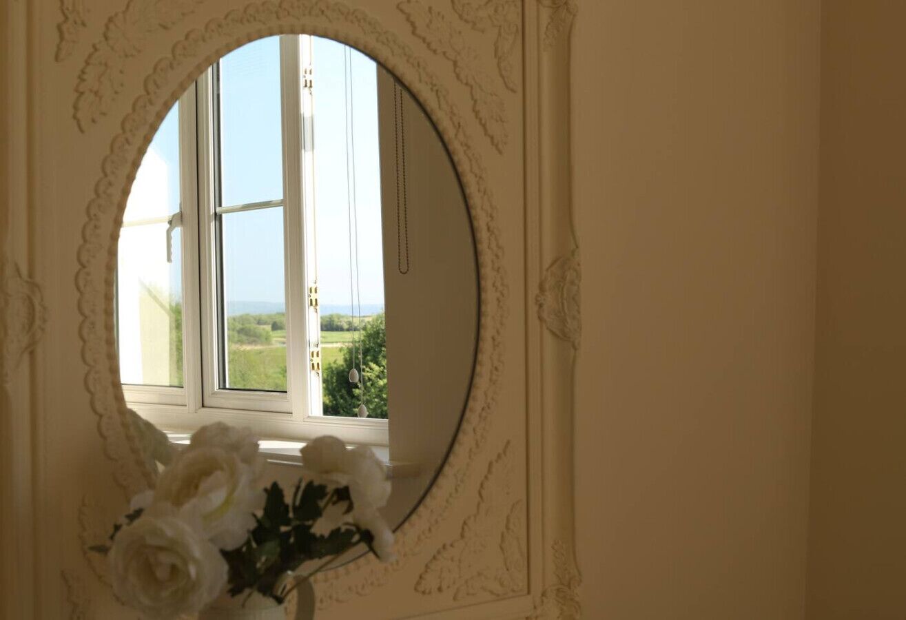 A room with a window framed by ornate molding, a view of greenery outside, and a vase with white flowers on a surface.