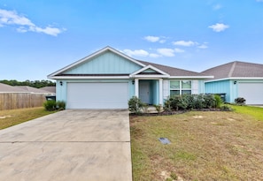 A single-story house with a garage, a front yard with grass and shrubs, and a clear blue sky. - Coastal Retreat Near Gulf Shores & Orange Beach (Foley)