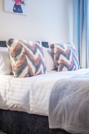 Ein Bett mit gemusterten Kissen, einer weißen Decke und einer grauen Wolldecke.