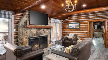 A log cabin interior with a stone fireplace, leather furniture, a mounted TV, and a chandelier.