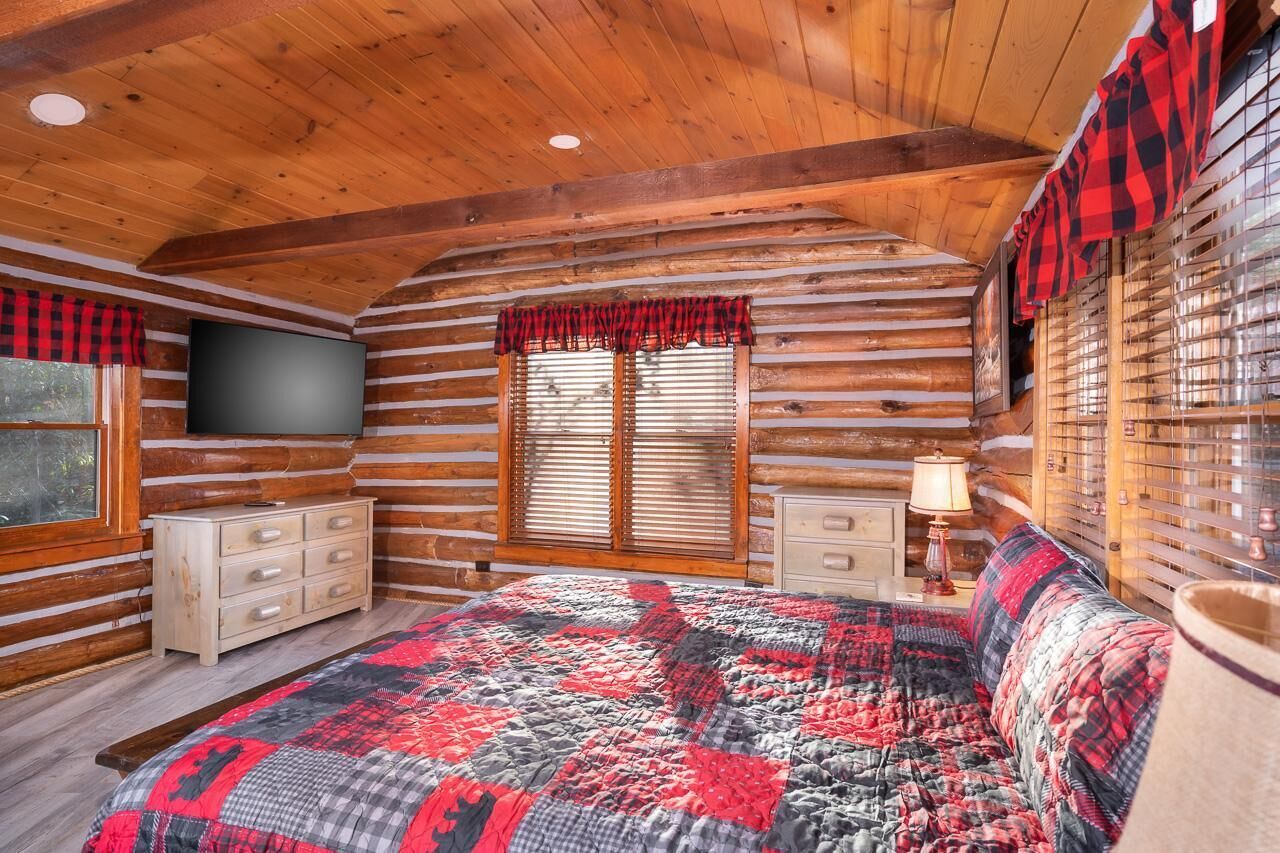 A log cabin bedroom with a bed, dresser, TV, and window with blinds.