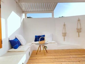 Une pièce baignée de lumière, avec un plancher en bois, des murs blancs, un banc agrémenté de coussins, une petite table sur laquelle est posé un panier, et des décorations murales en macramé.
