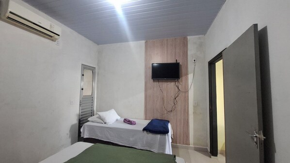 A small, single-bed room with a television mounted on the wall, a door, and an air conditioning unit.