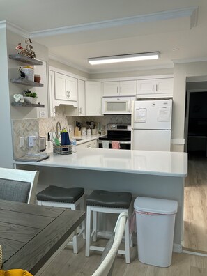 Eine moderne Küche mit weißen Schränken, einer zentralen Kochinsel inklusive Sitzgelegenheiten, einer Mikrowelle, einem Backofen und einem Kühlschrank.