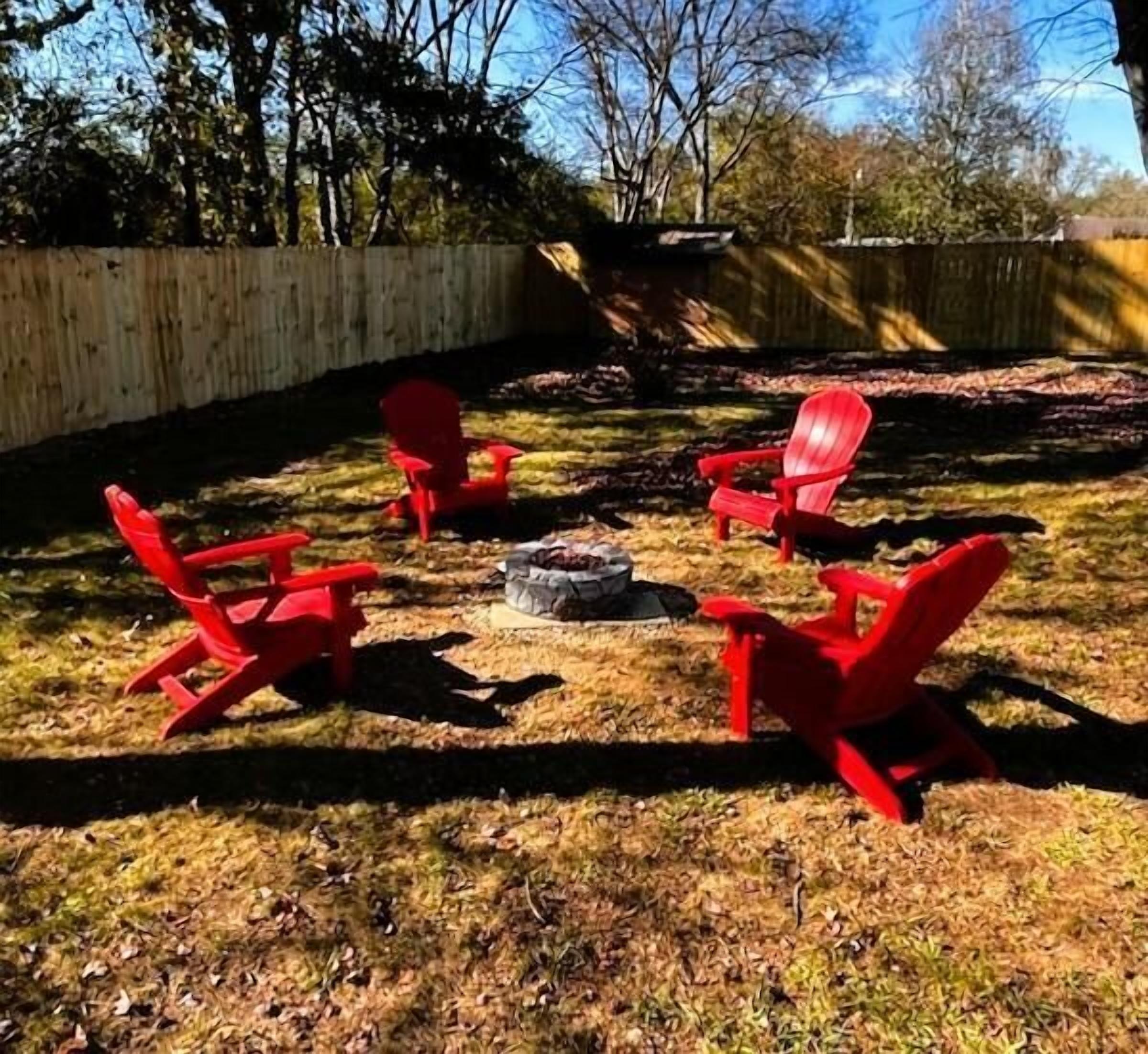 A backyard with a wooden fence, a fire pit, and four red Adirondack chairs arranged around it.