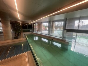 Una piscina interna con pareti di vetro trasparenti, pavimento piastrellato e vista sull'esterno.