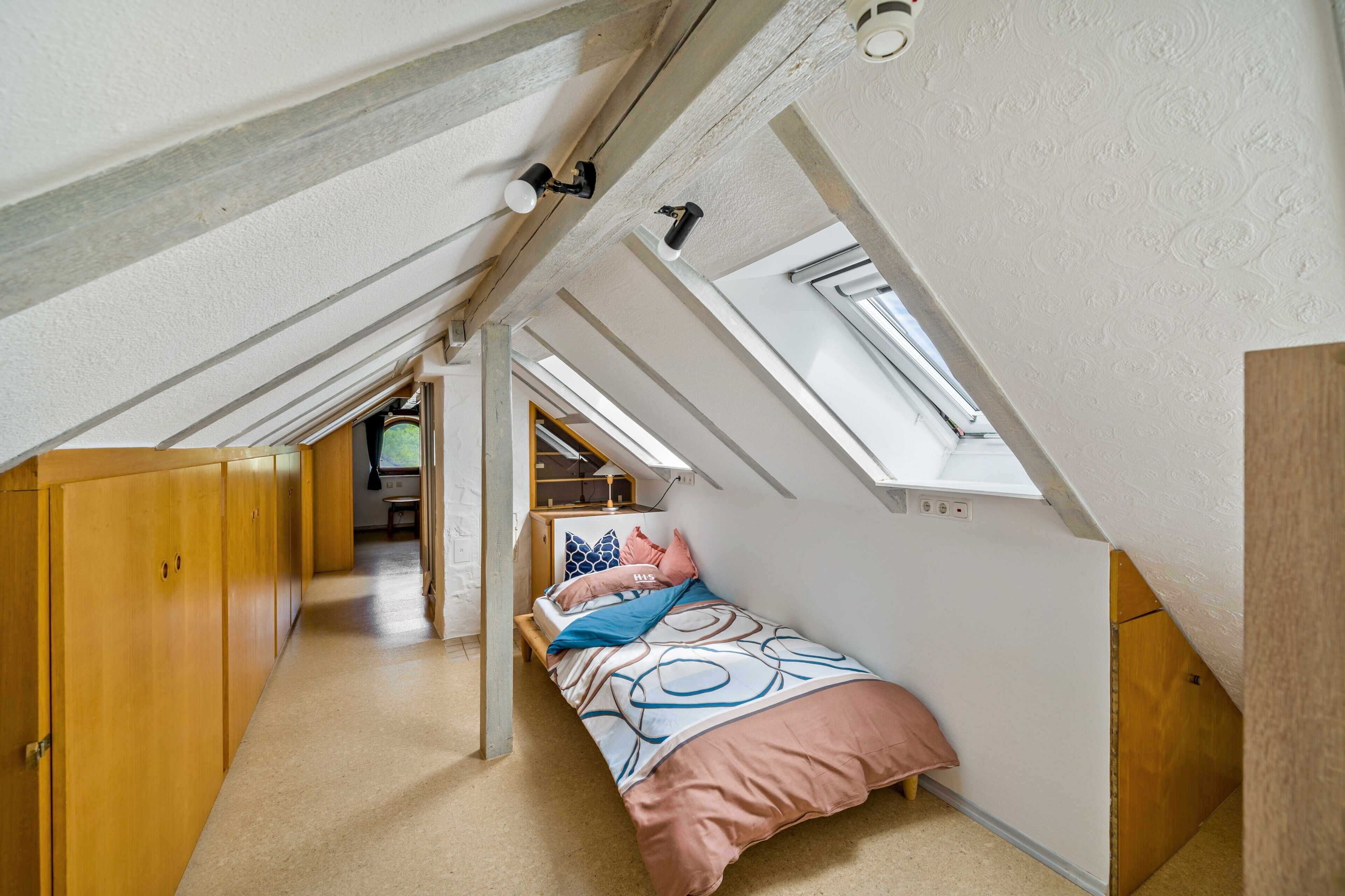 Ein kompakter Raum mit schrägen Wänden, einem Oberlicht, einem Holzschrank und einem Bett mit gemusterten Bettwaren.