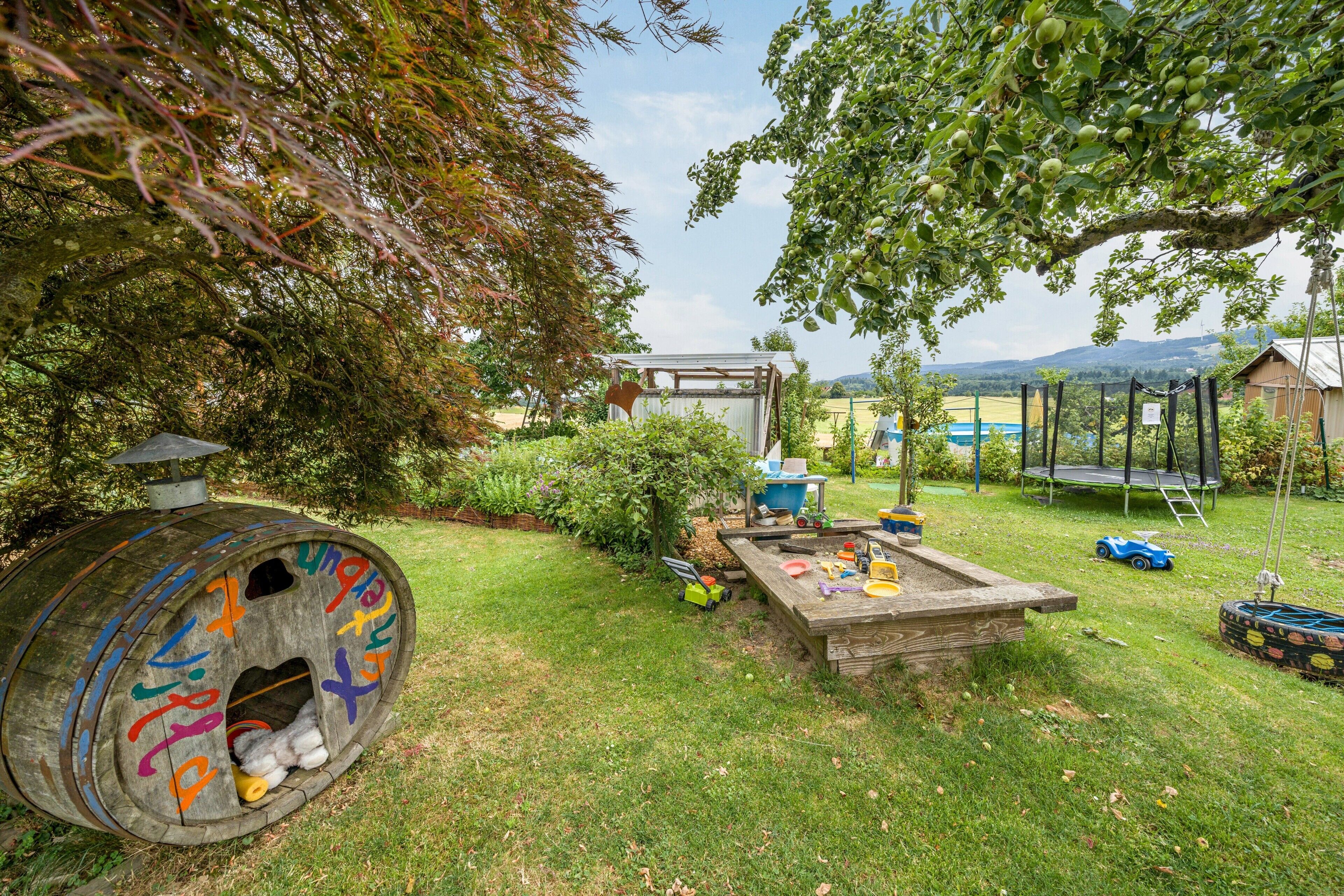 Ein Holzfass mit farbenfrohen Graffiti, ein Holztisch mit Spielzeug, ein Trampolin und eine Schaukel in einem grünen Hinterhof.