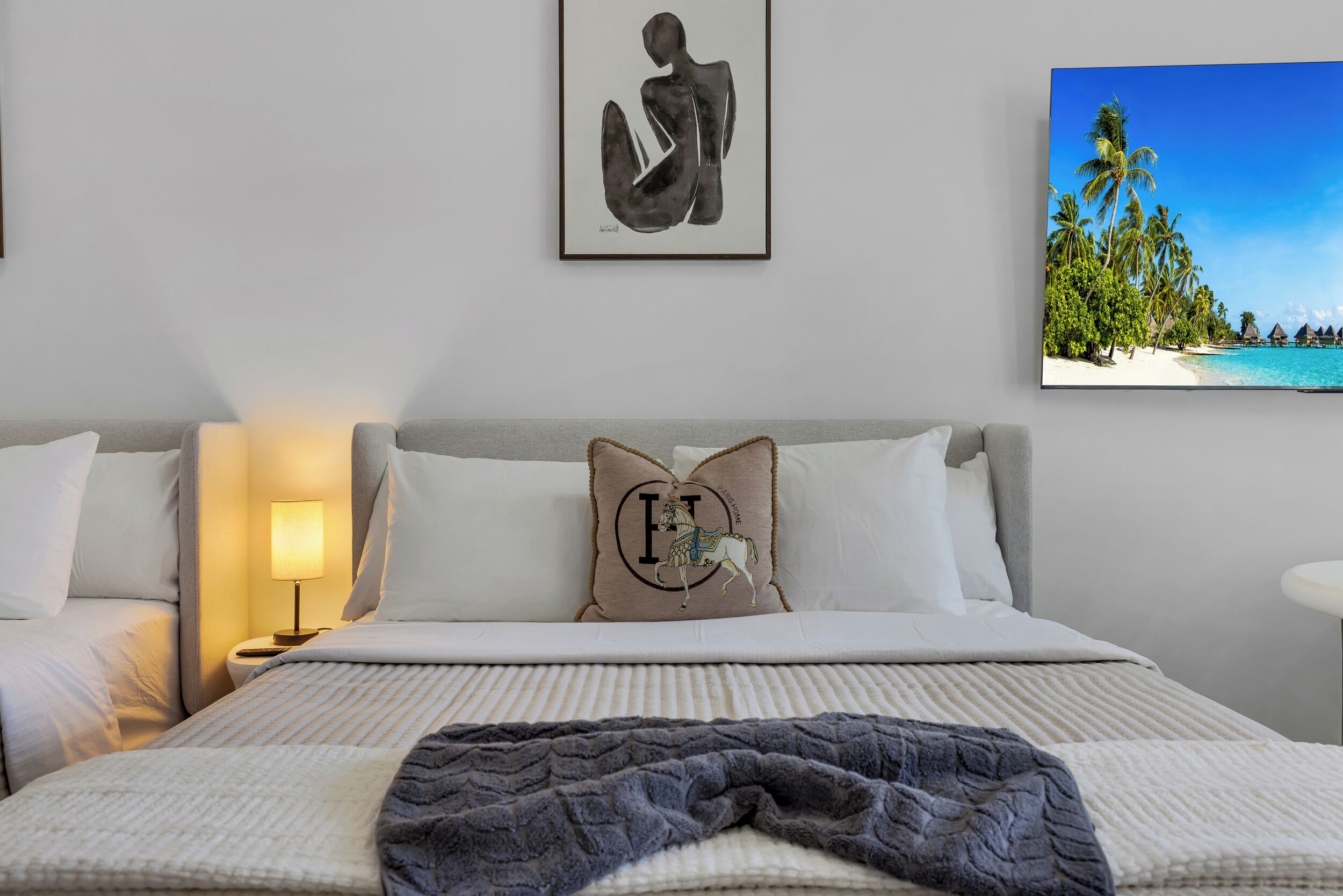 Una habitación de hotel con una cama, una mesita de noche, una lámpara, un almohadón con diseño de caballo y un cuadro enmarcado con una playa.
