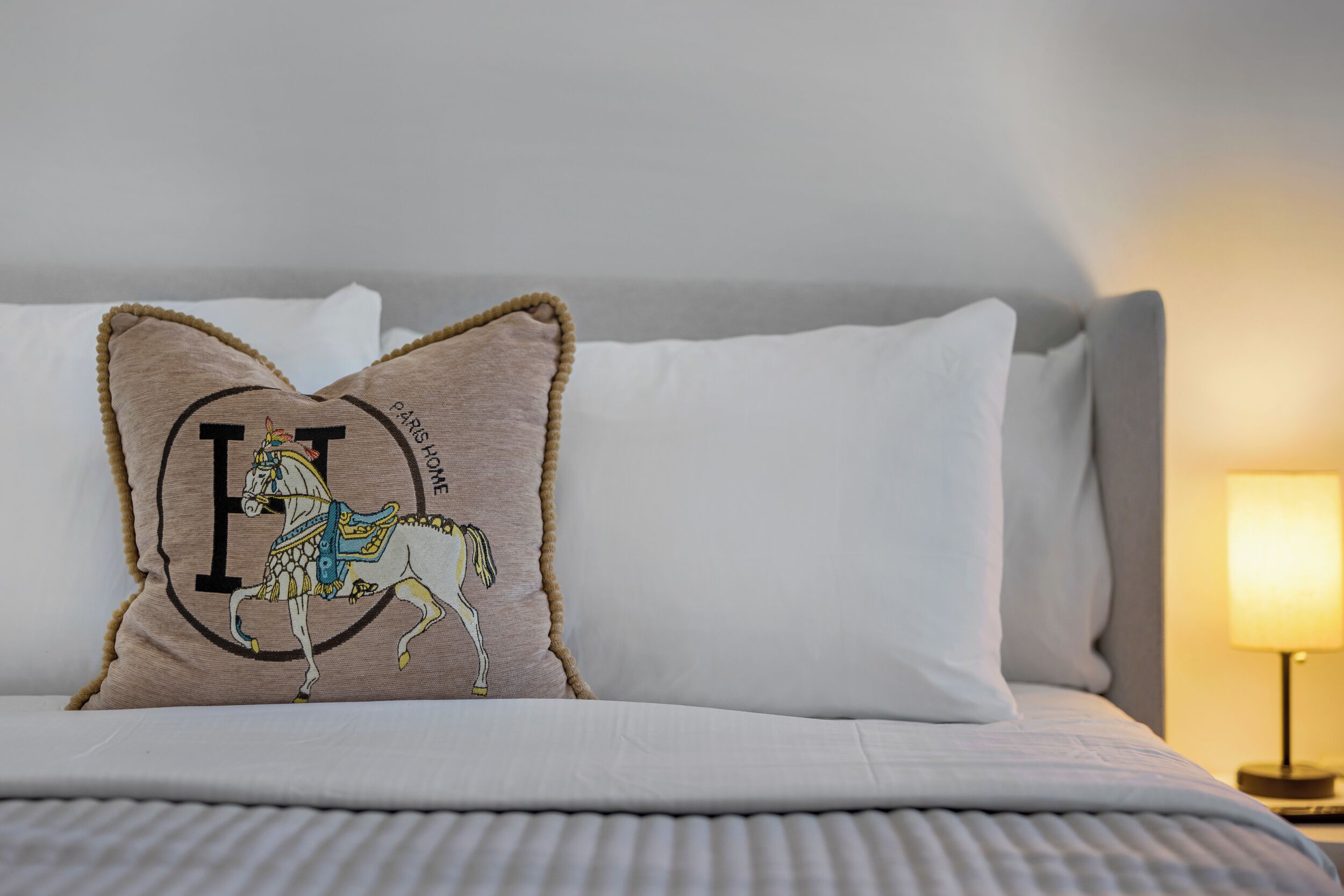 En seng med hvide sengetøj, en beige pude med hest og tekst 'PARIS HOME', samt en sengetøjslampe.