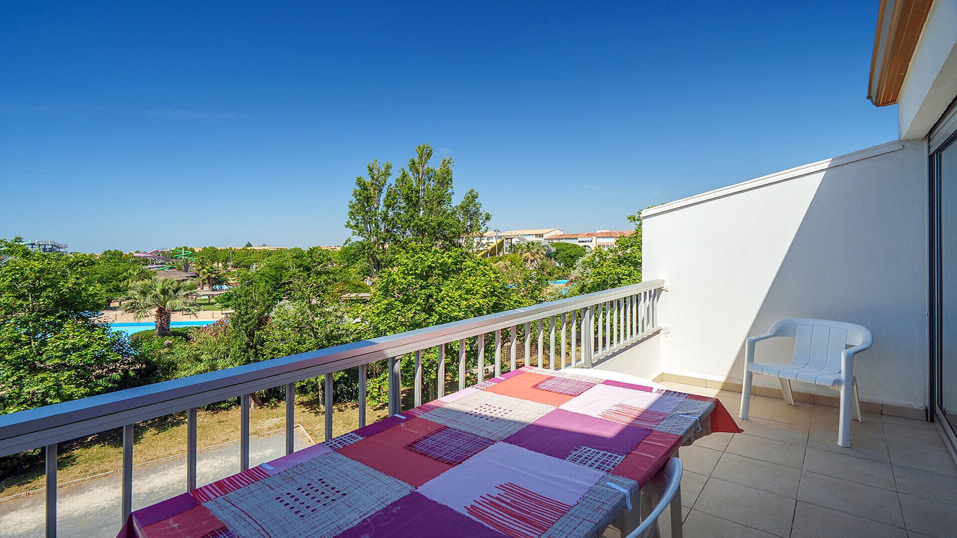 A balcony with a table covered in a patterned tablecloth, a white chair, and a view of a pool and buildings.