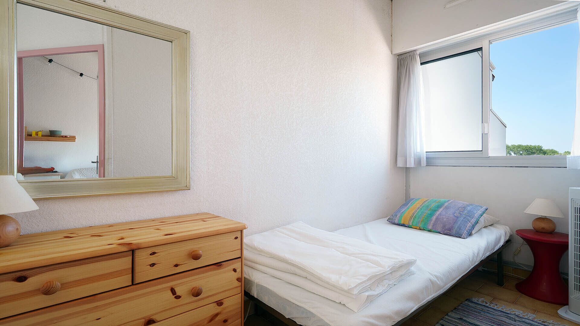 A bedroom with a bed, a dresser, a mirror, a window with curtains, and a small table with a lamp.