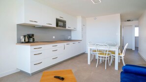 Eine moderne Küche mit weißen Schränken, einer eingebauten Mikrowelle und einer Kaffeemaschine. Im Wohnbereich befindet sich ein Esstisch mit weißen Stühlen und ein blaues Sofa.