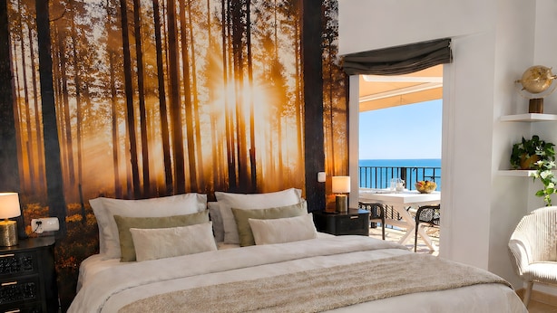 Ein Schlafzimmer mit Bett, Nachttischen, einer Lampe, Meerblick und einem Balkon mit Gartenmöbeln.