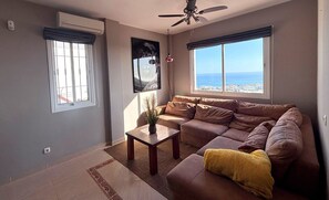 Ein Wohnzimmer mit einer Couch, einem Couchtisch, einem Deckenventilator und Blick aufs Meer.
