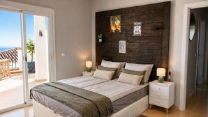 Ein Schlafzimmer mit einem großen Bett, zwei Nachttischen, einem kleinen Tisch am Bett und einem Holzkopfende, mit Blick ins Freie.