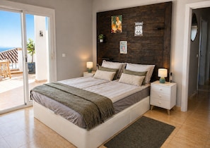 Ein Schlafzimmer mit einem großen Bett, zwei Nachttischen, einem kleinen Tisch am Bett und einem Holzkopfende, mit Blick ins Freie.