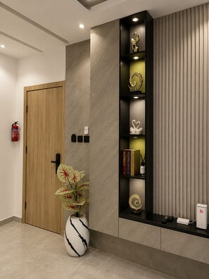 A modern reception area with a wooden door, a vase with a plant, a black shelf with decorative items, and a fire extinguisher.