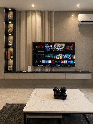 A modern living room with a flat-screen TV mounted on a wall, a coffee table, and a decorative shelf with books and ornaments.