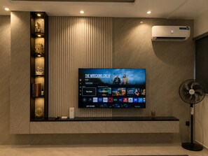 Un salon moderne dotĂ© dâun tĂ©lĂ©viseur Ă Ă©cran plat diffusant un film, dâun climatiseur intĂ©grĂ©, dâune bibliothĂšque agrĂ©mentĂ©e dâobjets dĂ©coratifs et dâun ventilateur sur pied.
