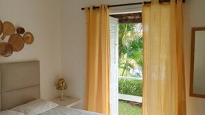 Une chambre à coucher avec un lit, un ventilateur de plafond, une fenêtre avec des rideaux et une vue sur la verdure à l’extérieur.